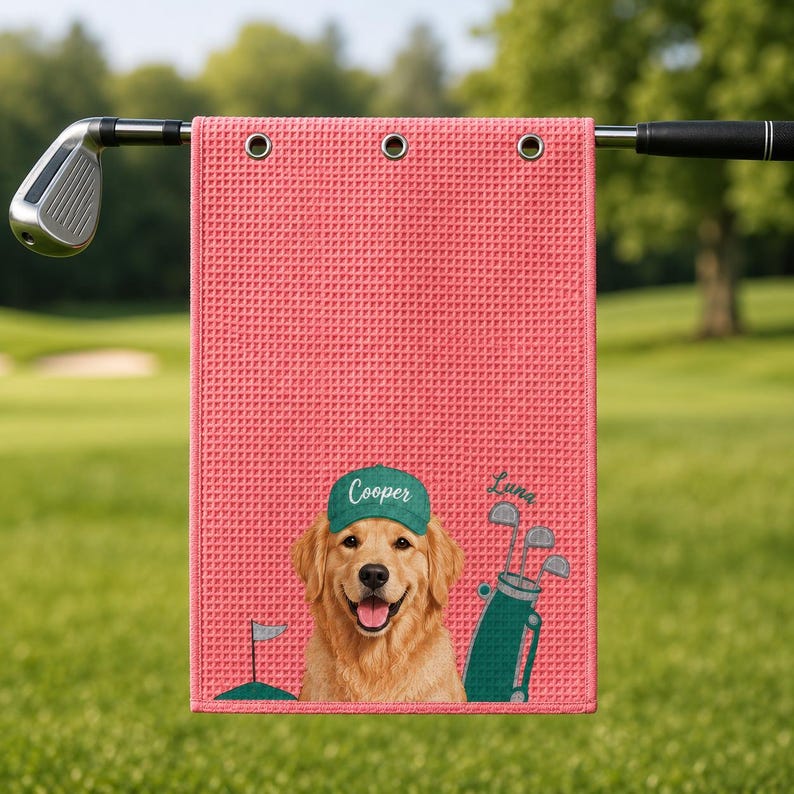 Puede incluir: Una toalla de golf color rosa coral con textura de panal que presenta un golden retriever con una gorra verde con el nombre Cooper impreso, junto a una bolsa de golf verde y el nombre Luna escrito en letra cursiva.