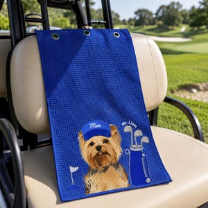 Puede incluir: Una toalla de golf azul real con textura de panal y tres ojales plateados en la parte superior. La toalla muestra un gr&aacute;fico de un Yorkshire Terrier con una gorra azul que dice Max, una bolsa de golf azul con palos y el texto Mr. Liam.