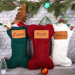 Könnte beinhalten: Drei Weihnachtsstrümpfe in Grün, Rot und Weiß, jeweils mit Zopfmuster. Der grüne Strumpf hat einen Rentierhut, der rote einen Zwergenhut und der weiße einen Zwergenhut. Jeder Strumpf hat ein Lederetikett mit einem Namen: Oscar, Pearl und Aaron.