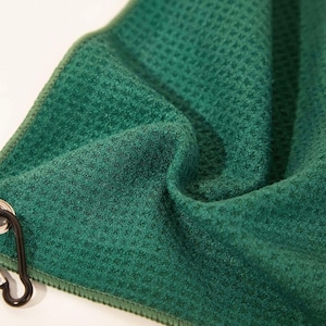 Puede incluir: Una toalla de color verde bosque profundo con un tejido de gofre y un patr&oacute;n de rejilla texturizado. La tela est&aacute; doblada para mostrar su profundidad y superficie suave. Un mosquet&oacute;n de metal negro est&aacute; unido a un ojal plateado en el borde.