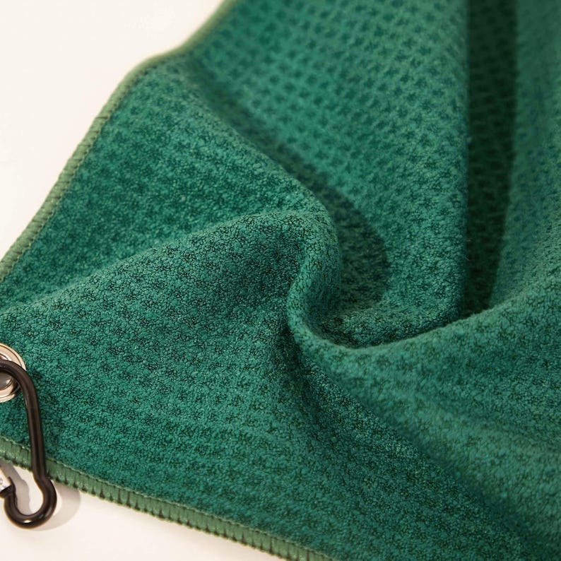 Puede incluir: Una toalla de color verde bosque profundo con un tejido de gofre y un patr&oacute;n de rejilla texturizado. La tela est&aacute; doblada para mostrar su profundidad y superficie suave. Un mosquet&oacute;n de metal negro est&aacute; unido a un ojal plateado en el borde.