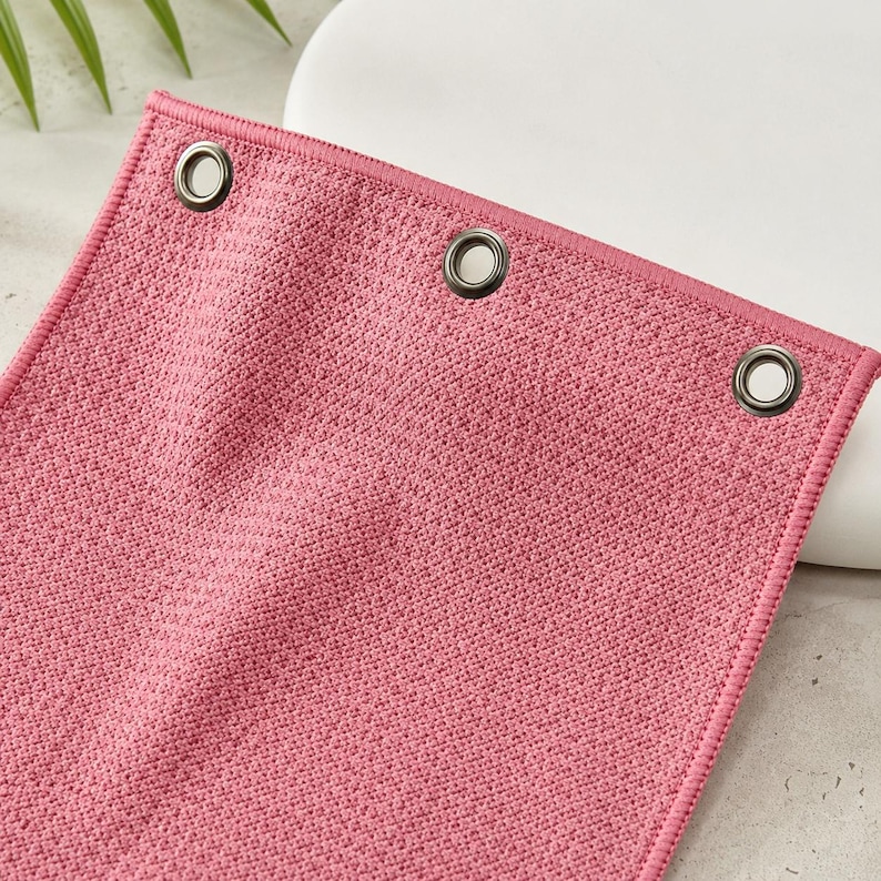Puede incluir: Una toalla rectangular de color rosa con un tejido texturizado tipo gofre. El borde superior presenta tres ojales met&aacute;licos oscuros espaciados uniformemente y reforzados con costuras. La tela tiene un color rosa s&oacute;lido y uniforme.