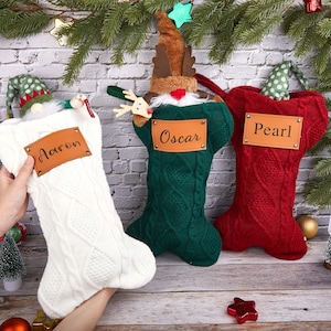 Op de afbeelding: Drie kerstsokken in wit, groen en rood, elk met een naamlabel. De witte sok heeft de naam "Aaron", de groene sok "Oscar" en de rode sok "Pearl". De sokken zijn versierd met feestelijke elementen.