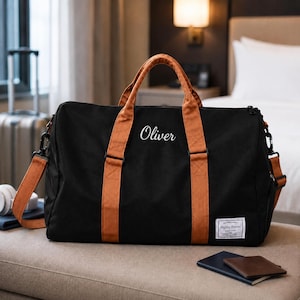May include: A black duffel bag with tan handles and a detachable shoulder strap. The bag features the name Oliver in white script lettering on the front and a small rectangular white label with the text Happy Travel in the bottom right corner.