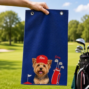Puede incluir: Una toalla de golf rectangular de color azul marino con textura de gofre, que presenta un gr&aacute;fico de un Yorkshire Terrier con una gorra roja con el nombre Max, una bolsa de golf roja con palos y el nombre Liam bordado en rojo.