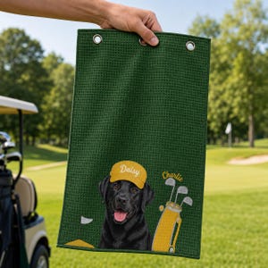 Puede incluir: Una toalla de golf de color verde oscuro con textura de gofre que presenta un labrador negro bordado con una gorra amarilla con el nombre Daisy, una bolsa de golf amarilla con el nombre Charlie y una peque&ntilde;a bandera de golf en un green amarillo.