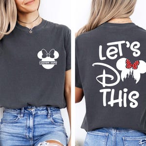 May include: Dark gray t-shirt featuring a white Minnie Mouse design with a bow and the text "CUSTOM NAME" on the front. The back of the shirt displays the text "Let's Do This" with a Minnie Mouse design and a red bow. The shirt is paired with blue denim shorts.