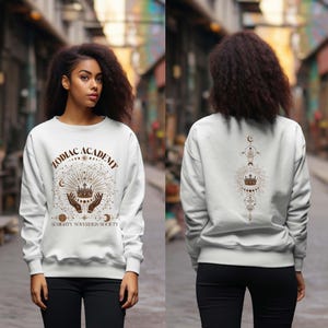 May include: White sweatshirt with a brown graphic print of a crown, hands, and celestial symbols. The text "Zodiac Academy Almighty Sovereign Society" is printed on the front of the sweatshirt.