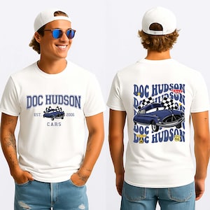 May include: White t-shirt with a blue cartoon car graphic and the text "DOC HUDSON" and "CARS" on the front. The back of the shirt repeats the graphic and text. The shirt also has the text "EST. 2006".