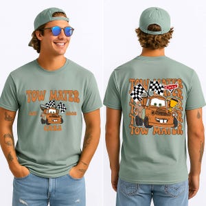 May include: Sage green t-shirt with a cartoon image of Tow Mater from the Cars film. The front of the shirt has the text "Tow Mater" and "Cars" with the year "2006". The back of the shirt has a larger image of Tow Mater with checkered flags.
