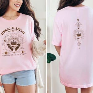 May include: Pink t-shirt with a brown graphic design featuring a crown, hands, and the text "Zodiac Academy Almighty Sovereign Society".