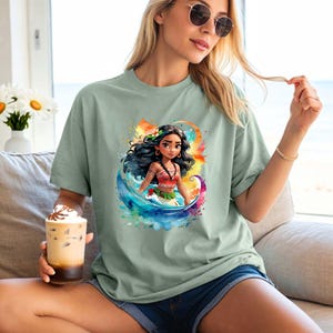 Peut inclure: Un t-shirt vert clair avec un motif aquarelle color&eacute; repr&eacute;sentant une princesse polyn&eacute;sienne de dessin anim&eacute; aux longs cheveux noirs, portant un haut rouge et orange et une fleur dans les cheveux. La princesse se tient dans une vague avec un fond de coucher de soleil.