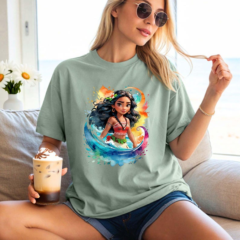 Peut inclure: Un t-shirt vert clair avec un motif aquarelle color&eacute; repr&eacute;sentant une princesse polyn&eacute;sienne de dessin anim&eacute; aux longs cheveux noirs, portant un haut rouge et orange et une fleur dans les cheveux. La princesse se tient dans une vague avec un fond de coucher de soleil.