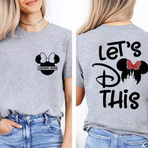 May include: Gray t-shirt featuring a black Minnie Mouse silhouette with a bow and the text "CUSTOM NAME" on the front. The back of the shirt has the text "Let's Do This" with a Minnie Mouse design.