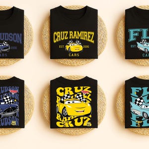 May include: Black t-shirts with car-themed graphics. Each shirt displays a different character's name, including "Doc Hudson," "Cruz Ramirez," and "Flo," alongside the year "2006" and the word "Cars."