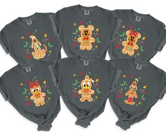 Op de afbeelding: Zes grijze t-shirts met opgerolde mouwen, elk met een peperkoekpersonage-ontwerp. De personages zijn Mickey Mouse, Minnie Mouse, Donald Duck, Katrien Duck, Pluto en Goofy, omringd door kerstversieringen.