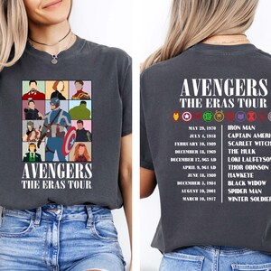 May include: A dark grey t-shirt with a graphic of the Avengers characters in a grid pattern. The text "Avengers The Eras Tour" is printed below the graphic. The back of the shirt has the text "Avengers The Eras Tour" printed at the top and a list of the Avengers characters with their corresponding dates.