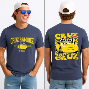 May include: Navy blue t-shirt with a yellow graphic of Cruz Ramirez from the Cars film. The front of the shirt says "CRUZ RAMIREZ EST. 2006 CARS". The back has a larger graphic of Cruz Ramirez with the text "CRUZ RAMIREZ".