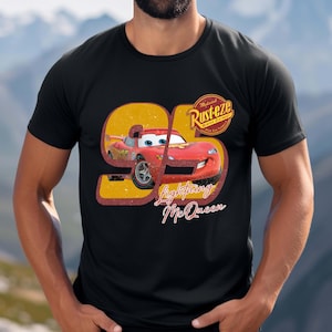 May include: Black t-shirt featuring a graphic of Lightning McQueen from the movie Cars. The design includes the number 95 in yellow and red, with the words "Lightning McQueen" in a cursive font. A "Rust-eze" logo is also present.