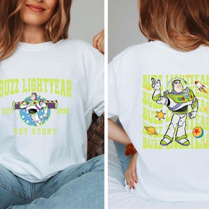 K&ouml;nnte beinhalten: Wei&szlig;es T-Shirt mit einem gr&uuml;nen und blauen Motiv von Buzz Lightyear aus Toy Story. Der Text "Buzz Lightyear" befindet sich in einem gelben Bogen &uuml;ber dem Motiv. Der Text "Est. 1995" befindet sich unter dem Motiv. Der Text "Toy Story" befindet sich unter dem Text "Est. 1995".