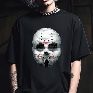 May include: Black t-shirt featuring a graphic of a hockey mask with a lake scene, a boat, and a figure with a weapon. The design is in shades of gray, red, and white. The shirt is worn by a person with silver hair.