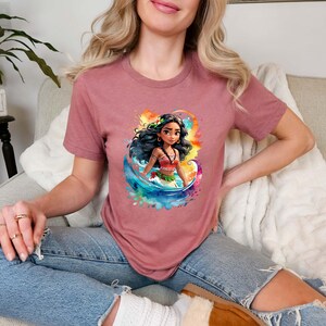 Peut inclure: Un t-shirt rose avec une image de dessin anim&eacute; d'une femme aux longs cheveux noirs, portant une tenue rouge et verte, debout devant une vague d'aquarelle bleue et verte. La femme porte une fleur dans les cheveux.