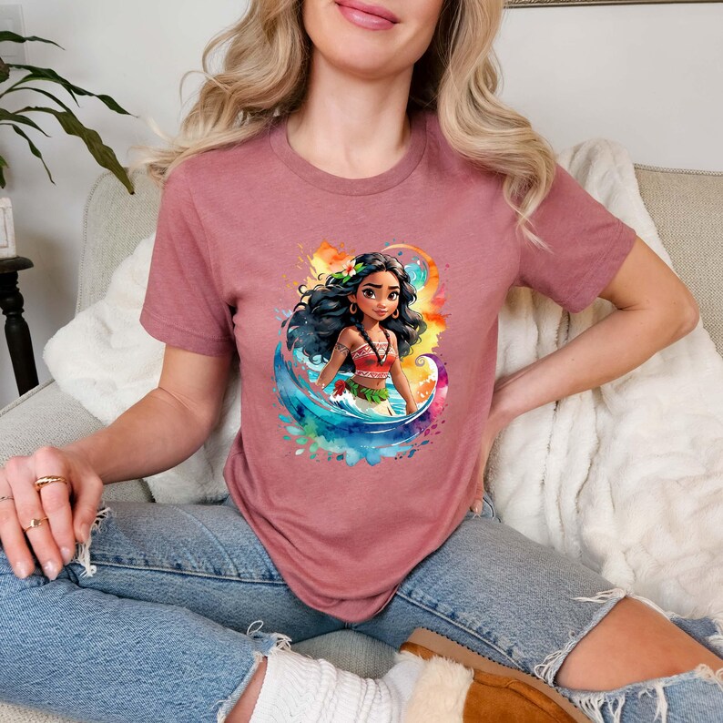Peut inclure: Un t-shirt rose avec une image de dessin anim&eacute; d'une femme aux longs cheveux noirs, portant une tenue rouge et verte, debout devant une vague d'aquarelle bleue et verte. La femme porte une fleur dans les cheveux.
