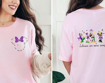 Puede incluir: Una camiseta rosa claro con un pequeño lazo morado y confeti de colores en la parte izquierda del pecho. La parte trasera muestra un gráfico de seis personajes de dibujos animados con ropa festiva y el texto Laissez les bons temps rouler.