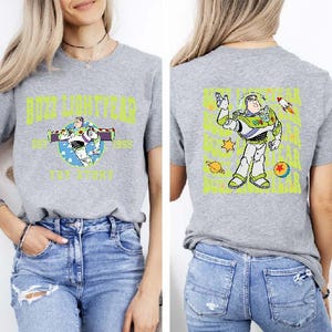 K&ouml;nnte beinhalten: Ein graues T-Shirt mit einer gr&uuml;n-blauen Comicfigur auf der R&uuml;ckseite. Die Figur ist Buzz Lightyear aus der Toy Story-Filmreihe. Die Vorderseite des Shirts zeigt eine Grafik von Buzz Lightyear mit dem aufgedruckten Text "Buzz Lightyear" und "Toy Story".