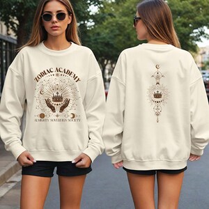 May include: A cream-colored sweatshirt with a graphic design on the front that says "Zodiac Academy Almighty Sovereign Society" with a crown and celestial symbols. The back of the sweatshirt has a similar design with a crown and celestial symbols.