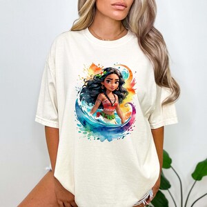 Peut inclure: Un t-shirt blanc avec un motif aquarelle color&eacute; d'une princesse polyn&eacute;sienne de dessin anim&eacute; aux longs cheveux noirs, portant un haut rouge et orange et une fleur dans les cheveux. La princesse se tient dans une vague avec un fond aquarelle bleu, vert et jaune.