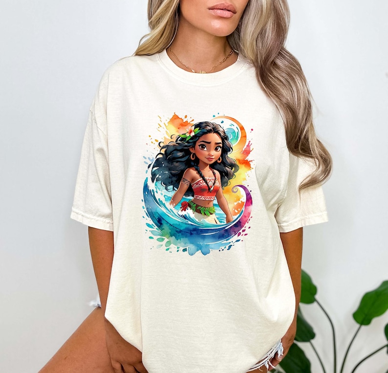 Peut inclure: Un t-shirt blanc avec un motif aquarelle color&eacute; d'une princesse polyn&eacute;sienne de dessin anim&eacute; aux longs cheveux noirs, portant un haut rouge et orange et une fleur dans les cheveux. La princesse se tient dans une vague avec un fond aquarelle bleu, vert et jaune.