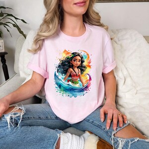 Peut inclure: Un t-shirt rose avec une illustration aquarelle color&eacute;e d'un personnage de dessin anim&eacute; aux longs cheveux noirs, portant un haut rouge et une fleur dans les cheveux. Le personnage se tient dans une vague.