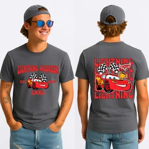 May include: Charcoal grey t-shirt featuring Lightning McQueen from the Cars film. The front graphic includes the text "Lightning McQueen" and "Cars", with a cartoon image of the car. The back graphic shows Lightning McQueen with checkered flags and a trophy.