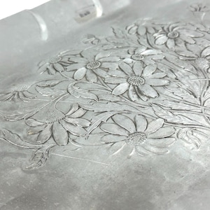 Może przedstawiać: Zbliżenie srebrnej tacy z kwiatowym wzorem. Taca ma wytłoczony wz&oacute;r stokrotek i liści. Szczeg&oacute;ły kwiat&oacute;w i liści są drobno oddane, tworząc teksturowaną powierzchnię. Taca ma kształt prostokąta.