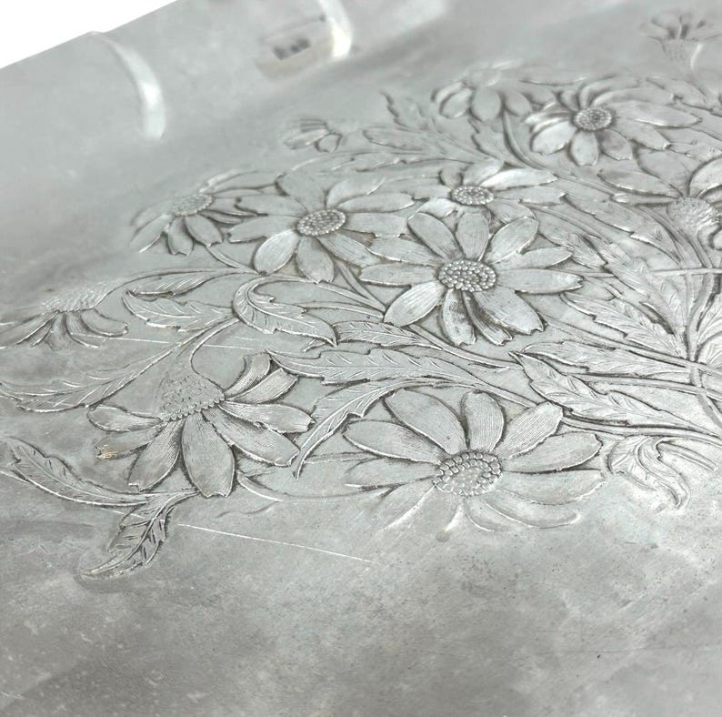 Może przedstawiać: Zbliżenie srebrnej tacy z kwiatowym wzorem. Taca ma wytłoczony wz&oacute;r stokrotek i liści. Szczeg&oacute;ły kwiat&oacute;w i liści są drobno oddane, tworząc teksturowaną powierzchnię. Taca ma kształt prostokąta.