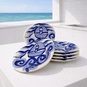 May include: Set of ceramic plates with a nautical theme. Each plate features a hand-painted blue sailboat design on a white background, surrounded by stylized blue waves. The plates are oval-shaped and stacked on a white surface.