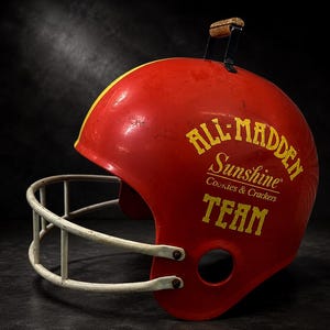 Puede incluir: Un casco de fútbol americano rojo con una franja amarilla y una máscara facial blanca. El lateral muestra el texto amarillo ALL-MADDEN Sunshine Cookies & Crackers TEAM. Un pequeño mango de madera está fijado en la parte superior.