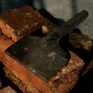 May include: A dark, weathered metal bricklayer's chisel rests on a pile of red-brown bricks. The chisel has a flat, rectangular blade and a long, rounded handle. The bricks are rough and textured, with some showing signs of wear.