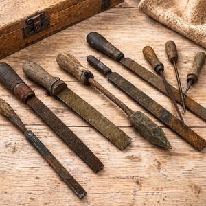 May include: A collection of vintage metal hand tools with weathered wooden handles, including various flat files, a pointed reamer, and small awls, arranged on a wooden surface next to a rustic wooden storage box with a metal latch.