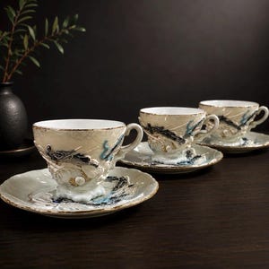 Op de afbeelding: Een set van drie crèmekleurige keramische theekopjes met bijpassende schoteltjes, voorzien van verhoogde, textuurrijke reliëfs van donkerblauwe en zwarte draken. Elk stuk heeft een delicate goudkleurige rand langs de geschulpte randen.