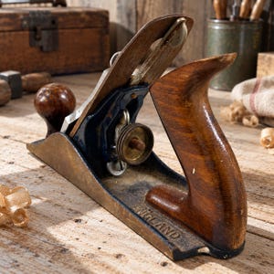 May include: A metal and wood hand plane featuring a dark blue body, a polished metal blade, and a brown wooden handle and knob. The side of the metal base is embossed with the word ENGLAND. The tool includes a circular brass adjustment dial.