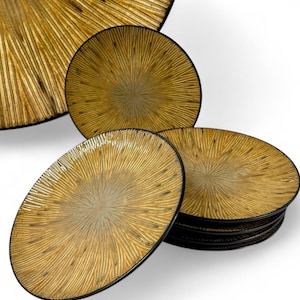May include: A set of gold-colored plates with a radial, textured design. The plates have a dark rim and a sunburst pattern emanating from the center. The plates are stacked and arranged to showcase the design.