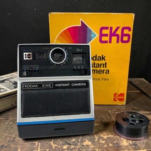 Puede incluir: Cámara instantánea Kodak EK6 vintage con cuerpo negro y plateado, detalles azules y el texto "KODAK EK6 INSTANT CAMERA". Una caja amarilla con "EK6" y "dak stant mera Print Film" está detrás de la cámara. También se ve un cartucho de película negro.