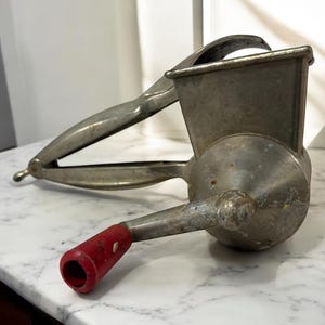 May include: Vintage metal food grinder with a red handle. The grinder has a silver finish and a lever-style handle. The design includes a hopper and a crank for operation. This kitchen tool is designed for grinding food.