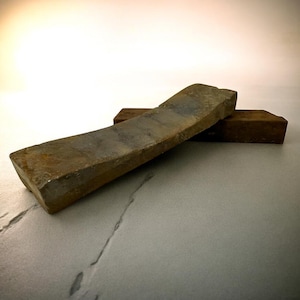 May include: Two sharpening stones. The larger stone is rectangular with a rough, textured surface and a grey-brown colour. The smaller stone is a dark brown colour and is positioned beneath the larger stone. The stones are on a light grey surface.