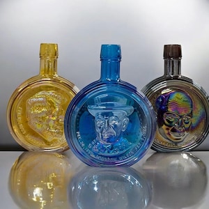 May include: Three decorative glass bottles in gold, blue, and iridescent colors. Each bottle features a portrait of a man in a hat. The bottles have a round shape with a flat front and a long neck.