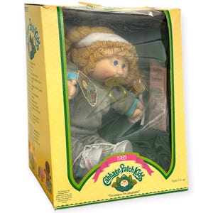 May include: A vintage Cabbage Patch Kids doll in its original box. The doll has blonde curly hair, blue eyes, and a pacifier. The box is yellow with green accents and features the Cabbage Patch Kids logo and the text "1985". The doll is suitable for ages 3 and up.