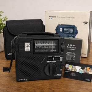 Könnte beinhalten: Ein schwarzes Eton FR-200 Notfall-Kurbelradio mit integrierter Taschenlampe, Lautsprechergitter und Abstimmknopf. Das Radio verfügt über eine Frequenzanzeige mit L.L. Bean-Branding, eine Handkurbel sowie die Originalverpackung, Anleitung und Tasche.