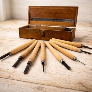 Może przedstawiać: Zestaw narzędzi do rzeźbienia w drewnie z drewnianymi uchwytami i metalowymi ostrzami, ułożonych na jasnej drewnianej powierzchni. W tle otwarta, zabytkowa drewniana skrzynka. Narzędzia mają różne kształty i rozmiary ostrzy.